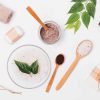 Flat lay arrangement of natural cosmetics for skin cleansing and exfoliation. Top view coffee scrub next to soap, sea salt, towel, pumice and green twig on white background.